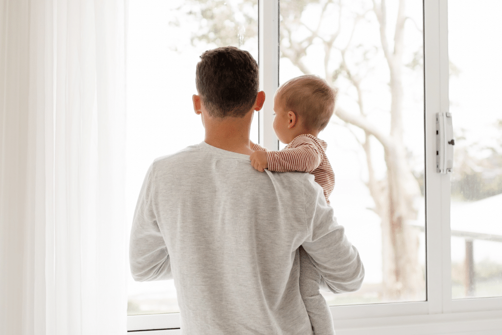 father and baby stand together with the back facing the camera. They are looking out the window