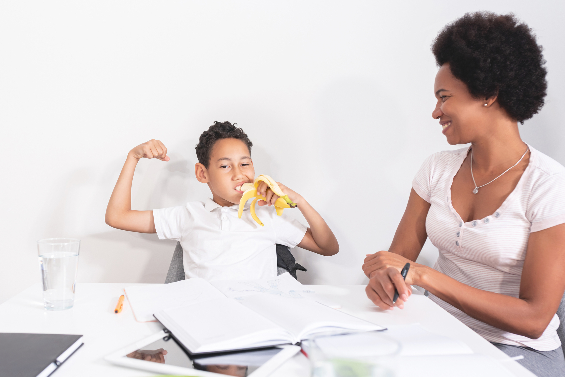 Tween appetite: Boy about age 12 eating banana while doing homework while mom looks on laughing. BIPOC family.