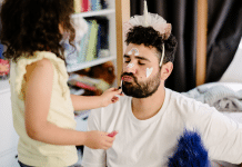 How Dads Are Updating The Script On Fatherhood A photo of fatherhood with daughter applies makeup to dad while playing dressup. The dad wears a unicorn crown and puckers up for lipstick