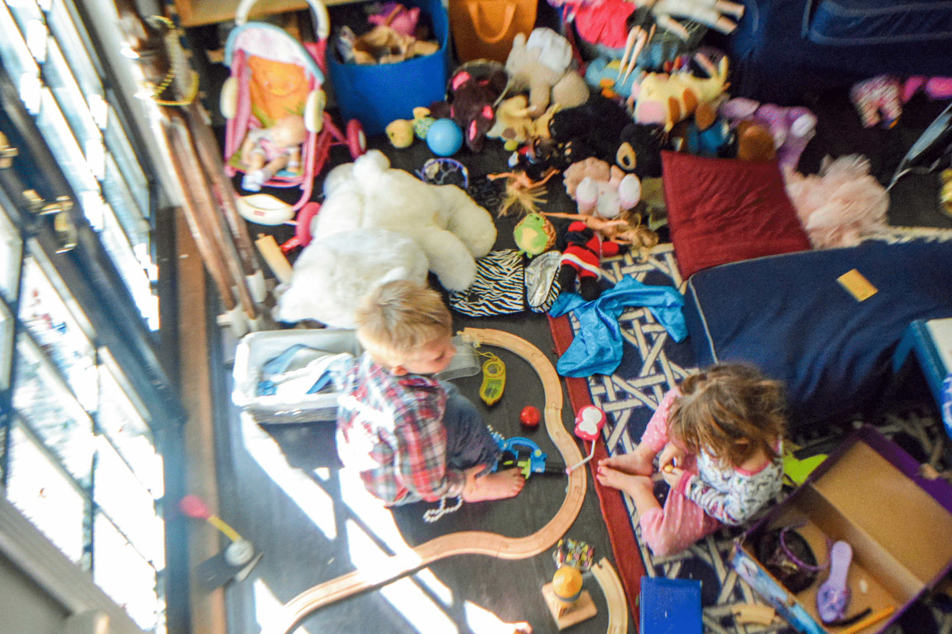 two children play on the floor of a very messy room with a floor covered in toys