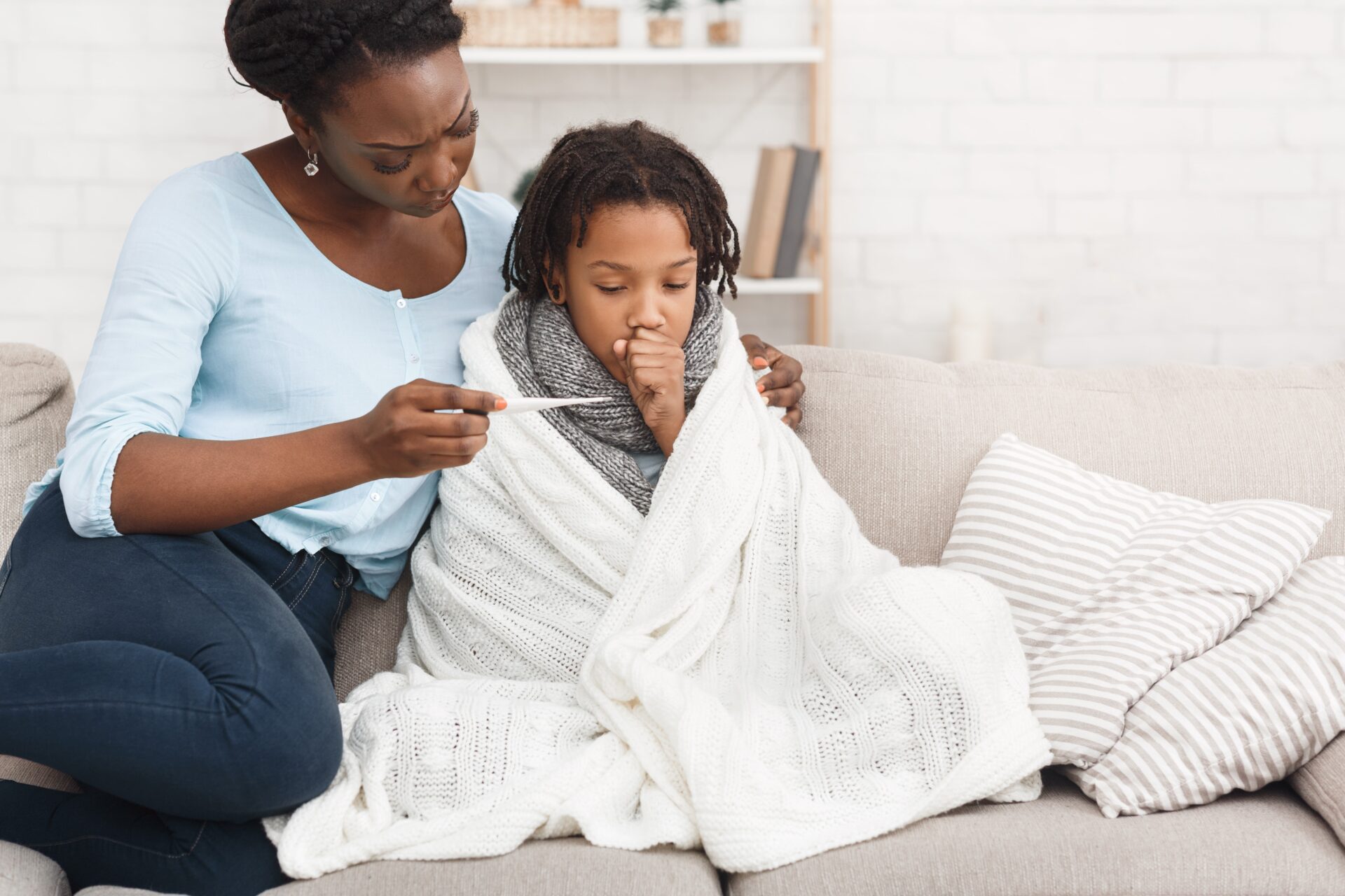 sick child with mom on the couch. Child is wrapped in a blanket with a thermometer