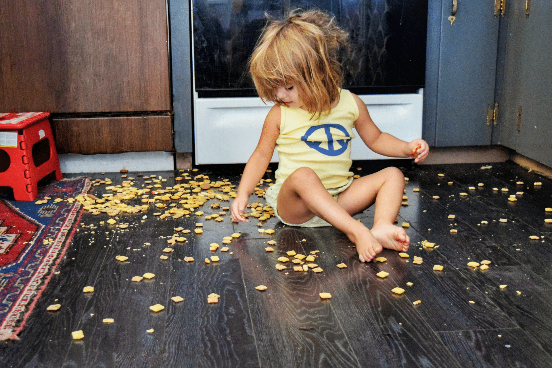 little girl sits on the kitchen floor with dumped crackers scattered all over