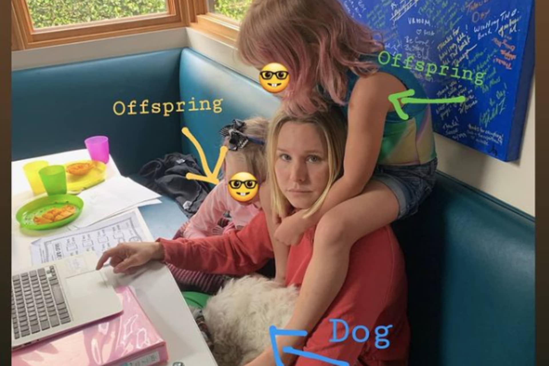 Kristen bell sits at the kitchen table looking frazzled with her dog and kids climbing on her