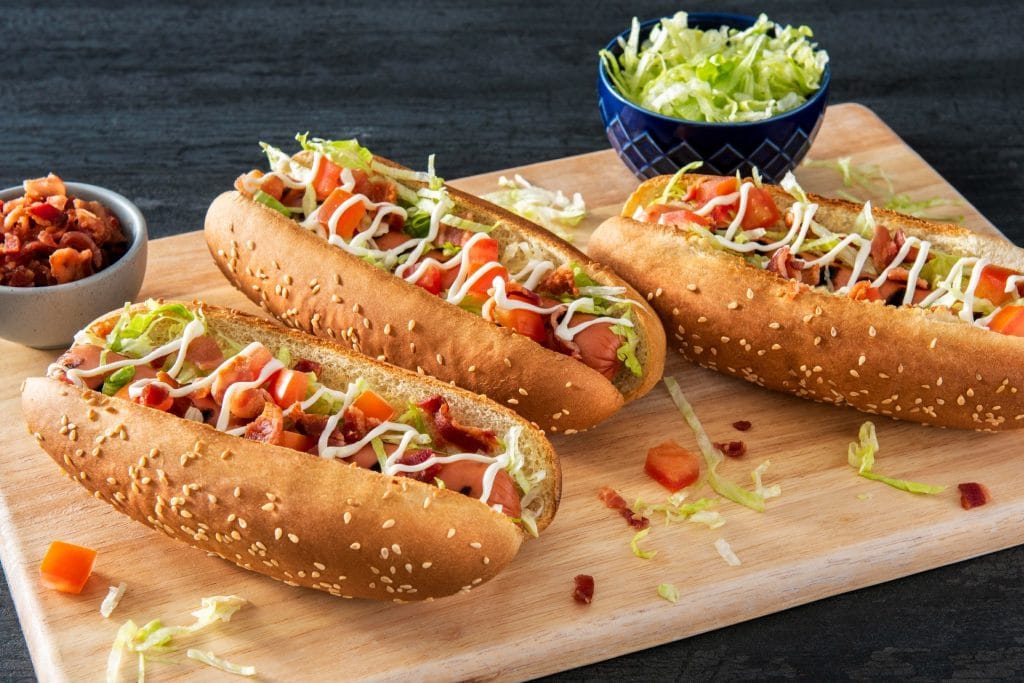 board with hot dogs topped with shredded lettuce, tomatoes and bacon