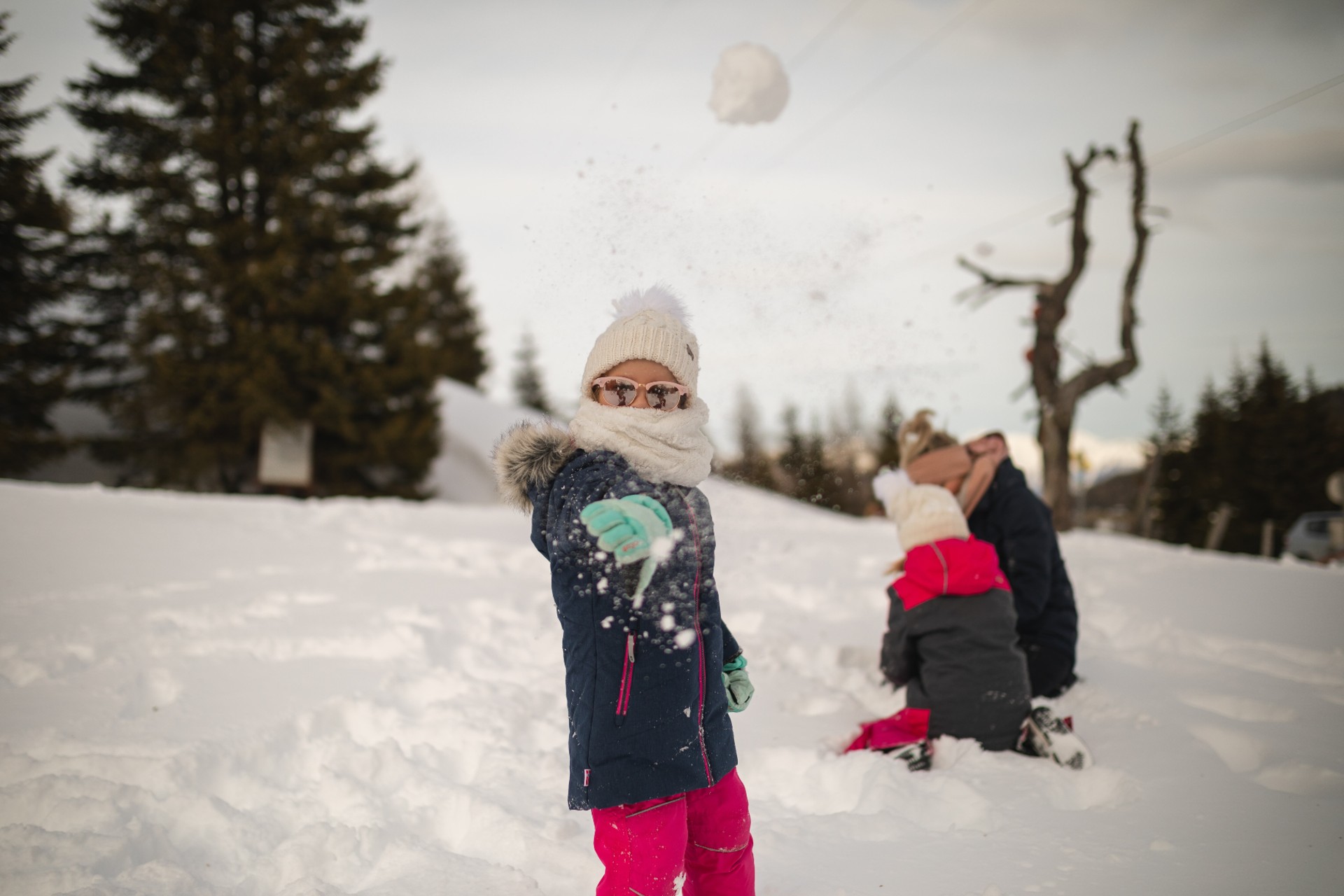 girl playfully throws a snowball towards the camera as her mom and sibling build a snowman in the background