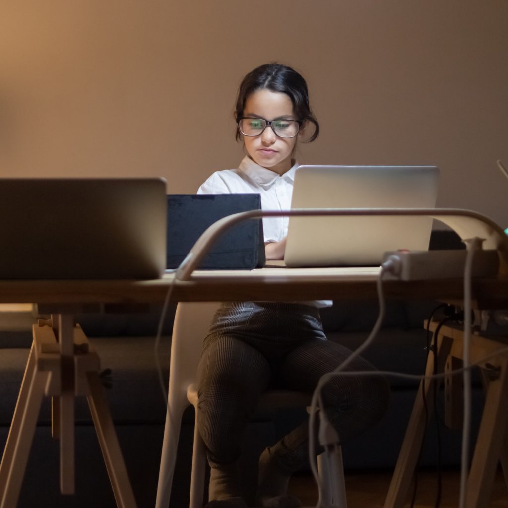 young teen girl with dark hair and dark glasses sits in dark room in front of multiple screens