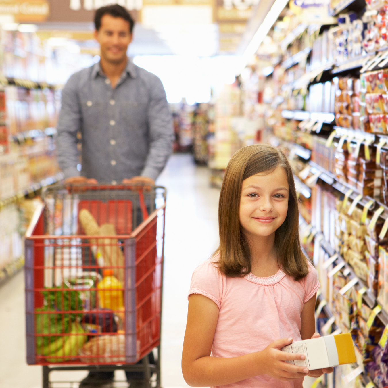 Male-presenting adult in background with female-presenting child in foreground at the grocery store. Adult is pushing a red cart filled with produce while the child smiles at the camera