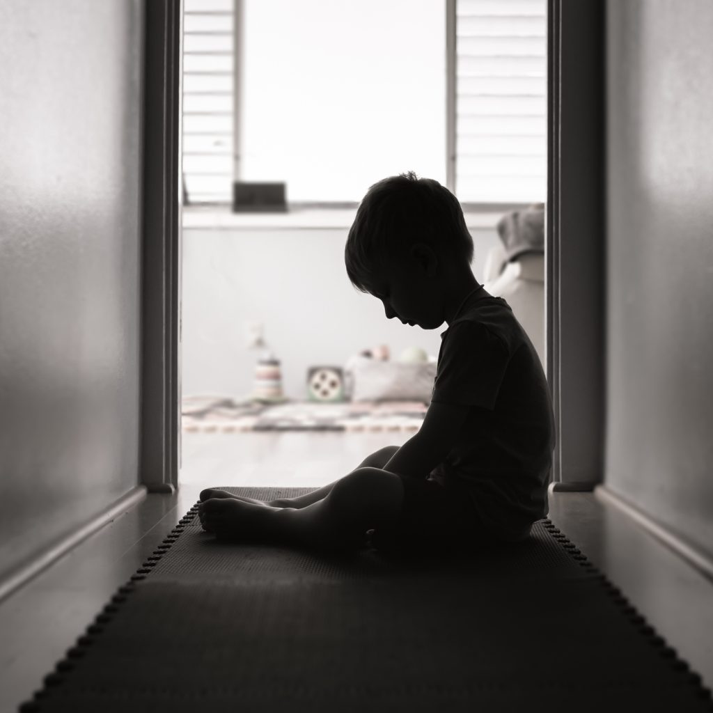 black-and-white image of child sitting in hallway looking down at feet and seeming very very sad