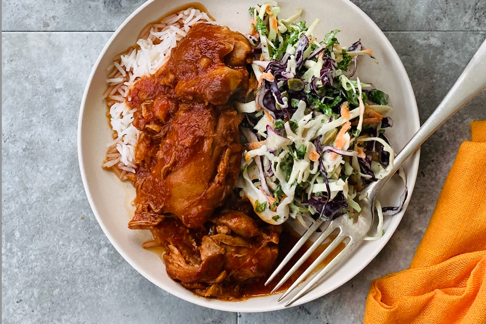 A plate of saucy chicken thighs on a plate with rice and slaw