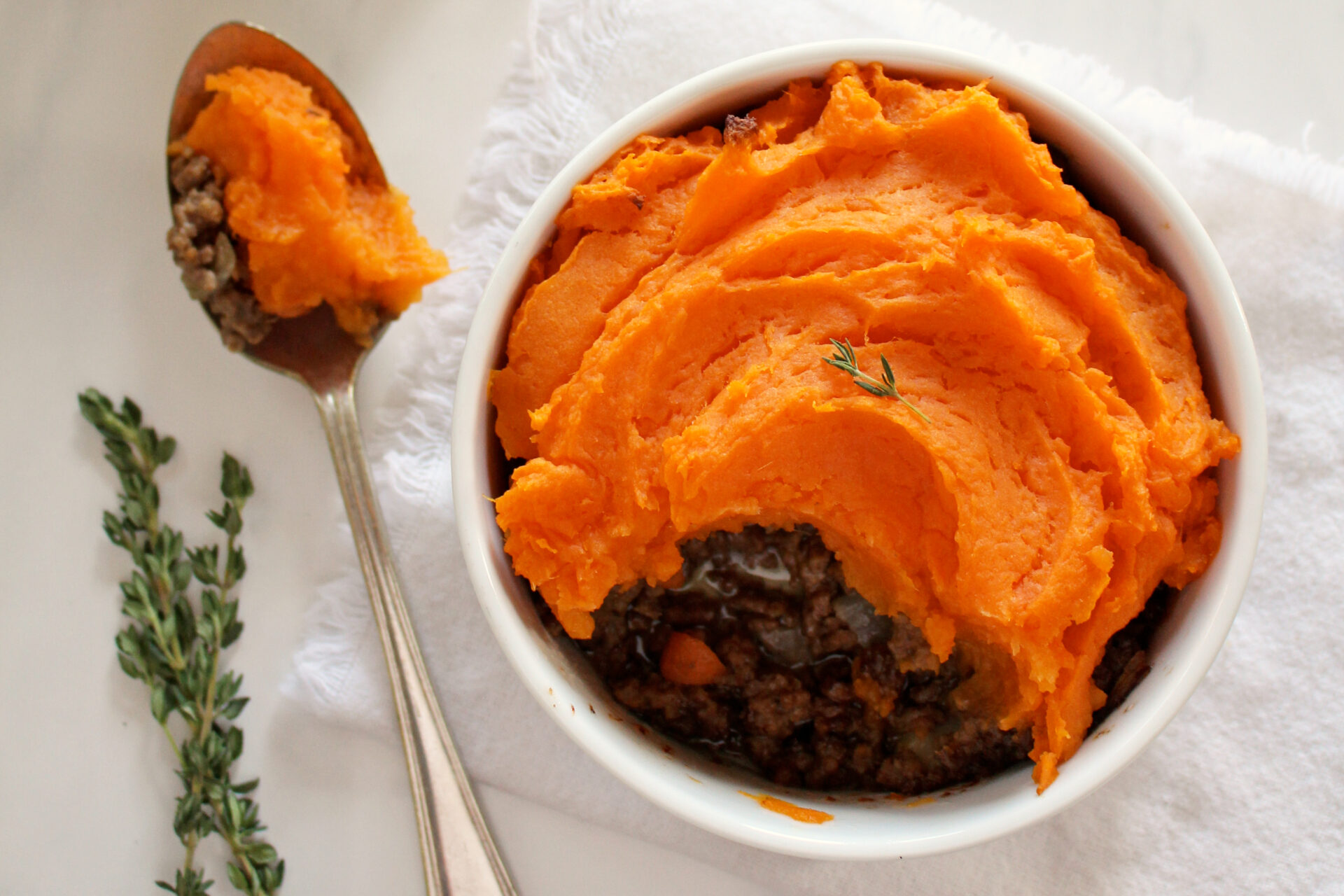 Bowl of sweet potato pie with bright orange mashed sweet potatoes covering beef mixture; one spoonful has been dug out to reveal bottom of pie and rests on spoon beside dish.