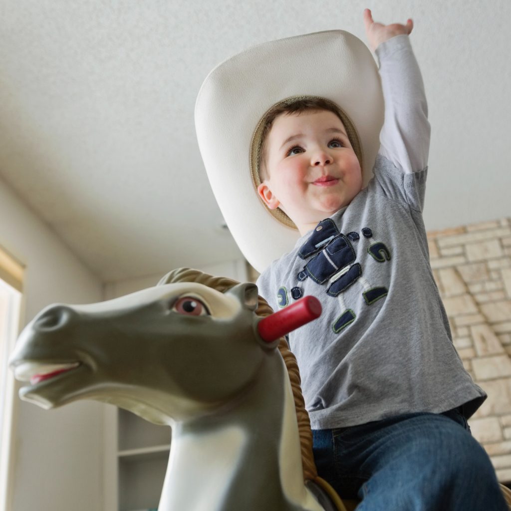 toddler boy in white cowboy hat rides a grey toy rocking horse