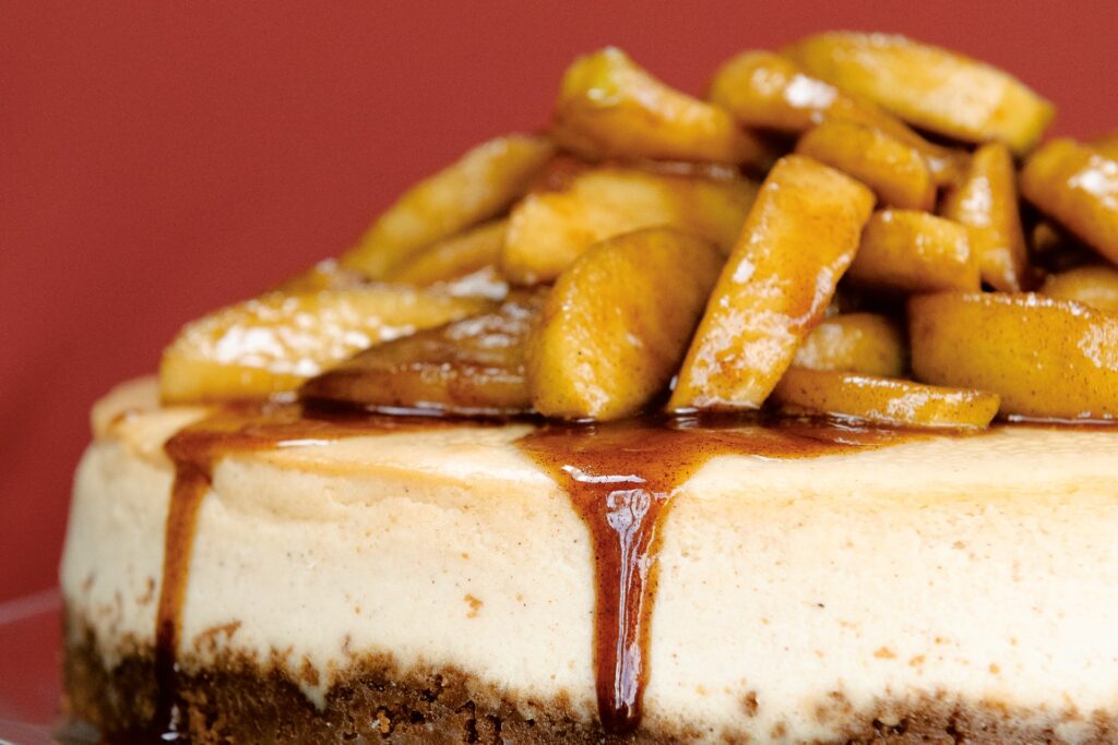 A spiced apple cheesecake sits on a pedestal with drippy caramel