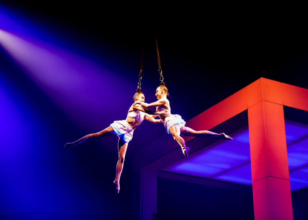 At Cirque Du Soleil: Echo, two acrobats hang in the air in stark blue light and a red fixture in the background.